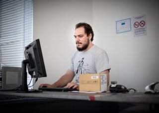 Un technicien travaille au clavier devant son écran, un colis posé sur le bureau de l’atelier.